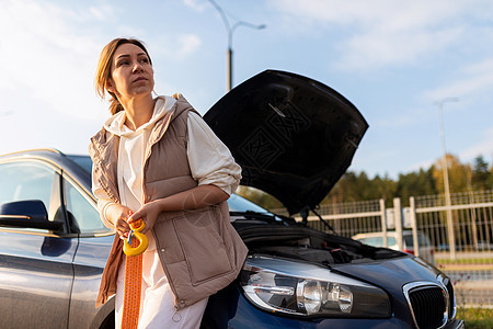 一名需要拖轮的妇女站在一辆破烂的汽车附近 车盖着一个开放的兜帽图片