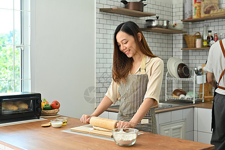 微笑的家庭主妇穿着围裙 在餐桌上用针卷面团 在现代厨房里做面包图片