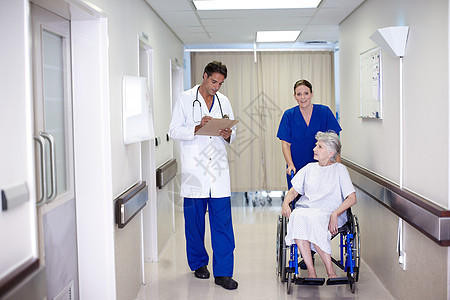 你做得很好 一位女护士在与她的医生交谈时 推着一位坐在轮椅上的老年患者走下走廊图片