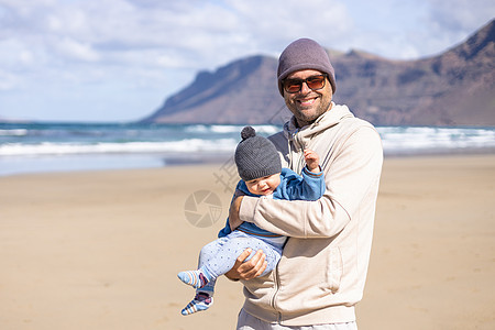 在西班牙兰萨罗特岛法马拉多风的沙滩上 父亲享受着纯净的大自然 抱着他的婴儿男婴儿子玩耍 家庭旅行和育儿理念图片