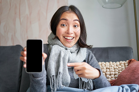 兴奋的年轻女性用手指指着智能手机 在手机上显示在线医生 医疗应用程序或 GP 联系方式 呆在家里生病 感冒药品流感症状成人长椅疼图片
