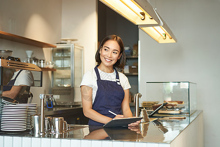 美丽的亚洲女孩笑着微笑的肖像 咖啡店的酒吧女招待在柜台后面工作 使用平板电脑作为POS终端 处理订单服务食堂药片女士命令咖啡食物图片