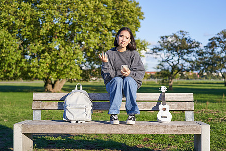 面脸悲伤 坐在长椅上与智能手机和ukulelele合唱机坐在一起的女孩乐趣成人微笑电话唱歌音乐家快乐女性工具弦琴图片