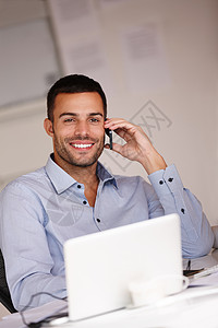 技术使他在比赛中处于领先地位 一个微笑的年轻人坐在他的办公桌前 正在使用他的手机图片