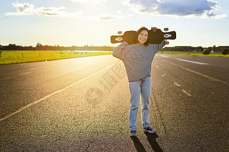 年轻的亚洲女性站在阳光明媚的路上 带着长板站在巡洋舰上的滑板公园滑冰青少年木板潮人女士街道青年活动滑冰滑板行动图片