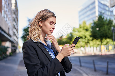 年轻女售货员的侧面肖像 穿着西装的公司女士 使用手机 在智能手机上阅读 站在办公室外的街道上面试营销成人自信金融企业家代理人电子图片