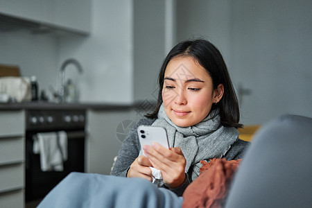 生病的韩国年轻女性坐在沙发上发短信 生病时用手机联系医生的画像病人长椅成人症状组织鼻子疼痛流感女士房子图片