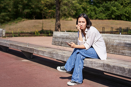 沮丧的亚洲女孩坐在公园里坐着长椅上 带着智能手机 感到不安和压力 惊慌和叹息治疗微笑喜悦电话福利学生享受黑发自由女性图片