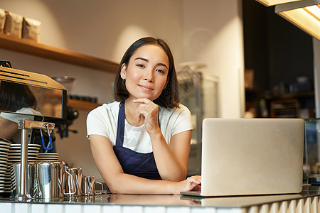 微笑的韩国女性的画像 咖啡店的咖啡师 拿着笔记本电脑站在柜台前 在她自己的咖啡店里微笑着 看起来自信 个体经营的女企业家人士服务图片