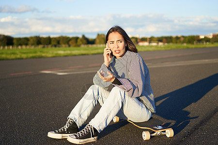 亚洲女孩的肖像坐在她的长板上 在滑板上滑冰 在手机上聊天 在电话交谈中发生冲突 与关心的脸争论潮人女性青少年青年乐趣街道公园运动图片