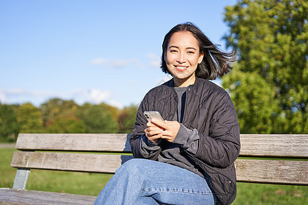 技术和人 使用智能手机的年轻女性 坐在户外公园的长椅上 看着智能手机 发送消息或联网成人幸福乐趣黑发爱好花园快乐女孩电话微笑图片