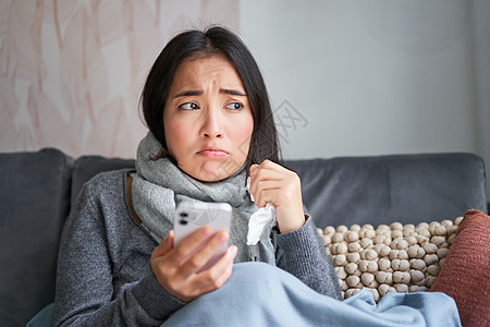 患有流感的亚洲女孩 坐在家里 哭泣 看起来很沮丧 拿着智能手机 感觉不舒服 阴沉 穿着暖和的衣服互联网组织成人女性过敏感染鼻子症图片