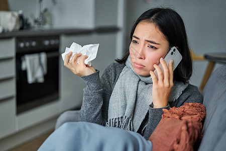 生病的年轻韩国妇女感到病痛 打喷嚏和拿着餐巾纸 呆在家里生病 感冒 在手机上交谈疼痛互联网疾病温度鼻子女士房子药品毯子过敏图片