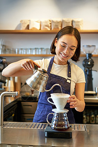 微笑的亚洲酒保 蓝色围裙的咖啡师 用小水壶倒水 在她的咖啡馆柜台后面冲泡咖啡的垂直照片工人玻璃职员食堂房子企业家女士商业酿造餐厅图片