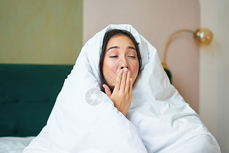 一个有趣的昏昏欲睡的女孩的特写 亚洲女人清晨醒来后打哈欠 用毯子盖住自己 手握在张开的嘴边床单房间女性寝具枕头小憩时间成人午睡就图片