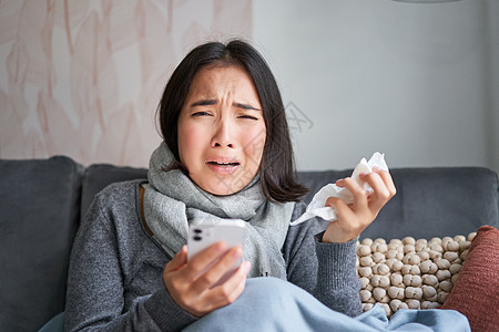 患有流感的亚洲女孩 坐在家里 哭泣 看起来很沮丧 拿着智能手机 感觉不舒服 阴沉 穿着暖和的衣服互联网温度药品毯子疼痛病人发烧感图片