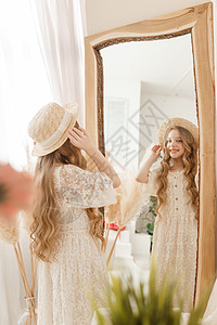 长发的美少女在镜子前戴上草帽 对金发女郎的自以为是的感觉幸福裙子女孩身体衣服拥抱头发帽子稻草装饰图片