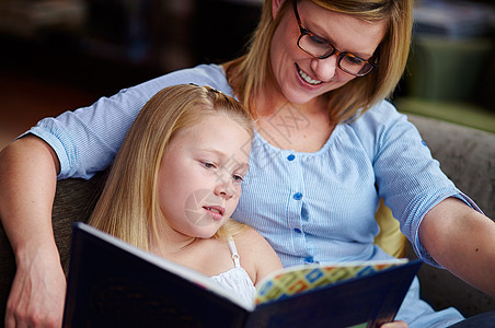 一位可爱的年轻女孩坐在她母亲旁边看书时 当时他们正在阅读一本书图片