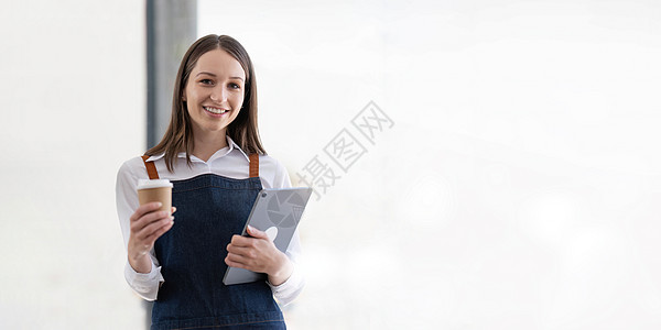 美丽的亚洲年轻咖啡师女人穿着围裙拿着平板电脑和杯子站在咖啡馆门前 上面有开放的招牌 企业主创业中小企业企业家概念图片