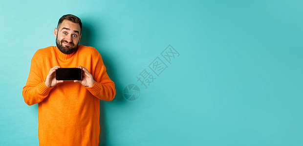 无能的人耸耸肩 展示手机屏幕 犹豫不决的情绪 站在绿松石色背景上的橙色毛衣购物黑发快乐潮人橙子促销喜悦标识横幅男性图片