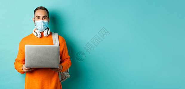 年轻男子用笔记本电脑戴面罩 对着浅蓝色背景站立的照相机欢笑地凝视着隔离毛衣快乐胡子冒充社交情感橙子购物手势图片