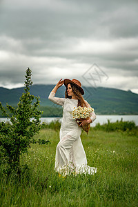 一位身穿白色连衣裙和棕色帽子的中年妇女手里拿着一个篮子 上面放着一大束雏菊 祝贺的野花乡村植物甘菊草地季节荒野植物群洋甘菊天空花图片