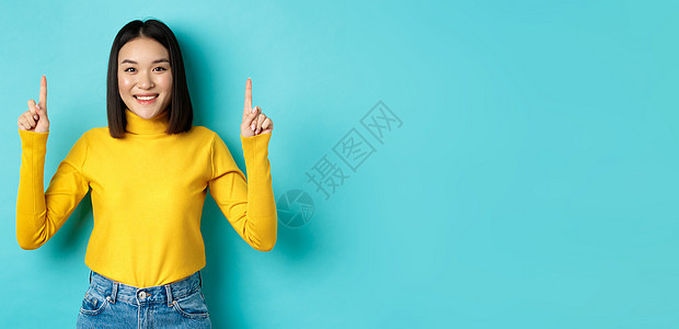 购物概念 美丽的穿黄色毛衣的Vietnames女性形象 展示广告 微笑和指着手指站立在蓝底的背景上黑发手势女孩工作室发型青少年成图片