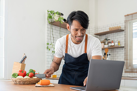 厨房里的男人在做饭时看笔记本电脑上的食谱营养早餐成人围裙饮食桌子休闲装厨师沙拉电话图片