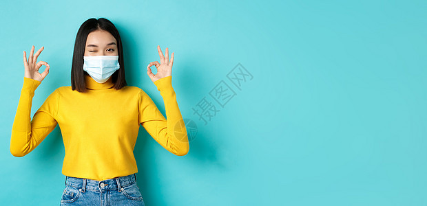 Covid19 社会距离和流行病概念 戴着医用面具的快乐亚洲女孩表现出良好的手势 对着镜头眨眼 表示赞同 站在蓝色背景上交易药品图片