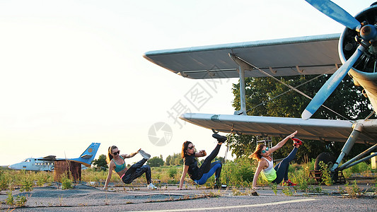 戴着墨镜 穿着紧身衣 漂亮 运动的年轻女性同步进行不同的力量练习 跳跃 俯卧撑 抬腿 在飞机附近的废弃机场杠铃耐力活动男性培训师图片