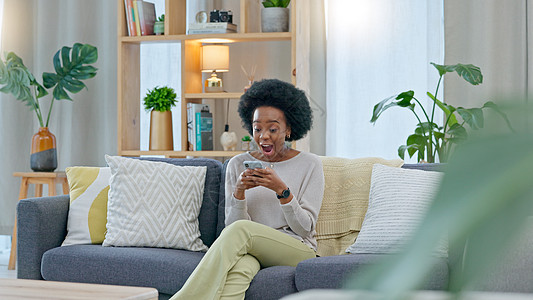 非洲妇女坐在家里的沙发上庆祝一份新工作 一位年轻女性的贷款通过她手机上的电子邮件获得批准 一位快乐而兴奋的女士在沙发上为晋升而欢图片