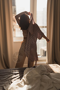 年轻妇女在卧室享受阳光明媚的早晨     觉醒和健康概念喜悦房子窗帘太阳窗户公寓睡衣晴天活力唤醒图片