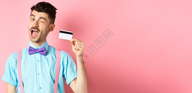 购物概念 有趣的家伙 胡子的胡须在镜头上眨眼 展示塑料信用卡 建议银行宣传出价 站在粉红色背景上成功金融微笑促销吊带裤广告发型商背景图片