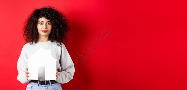 房地产 穿着休闲服的年轻白人女性站在红色背景上展示纸屋剪纸 购买房产或租房抵押卷曲发型机构女孩广告保险女士房子生活图片