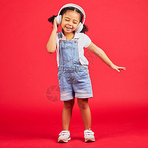 在孤立的红色背景下 戴着音乐耳机 有趣的收音机或儿童播客跳舞 精力充沛或小女孩 微笑 快乐或跳舞的孩子在工作室模型上听音频 声音图片