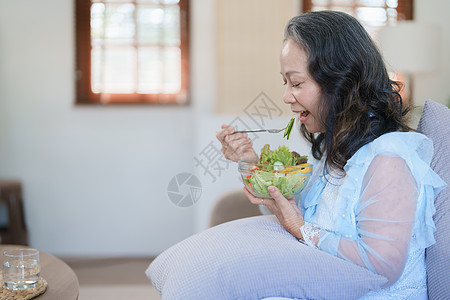 一位年长的亚洲妇女通过吃沙拉来照顾自己的健康水果祖母沙发微笑老年午餐桌子退休夫妻成人图片
