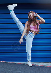 城市蓝色背景中疯狂的音乐 自由和兴奋的女孩在耳机上听曲目和音频 时尚 生活方式和为跳舞 快乐的态度或流媒体歌曲而抬腿的女人图片
