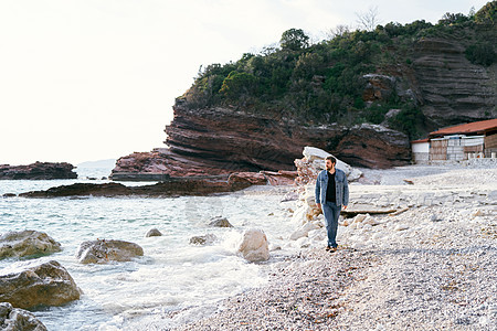 穿牛仔裤和戴尼姆夹克的五肢男子在一块石头沙滩上行走 以绿色岩石为背景看着水图片