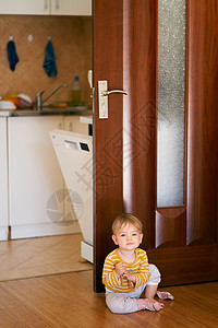 穿着条纹衬衫的可爱孩子坐在厨房地板上 背景是开着的门和后面的洗碗机图片