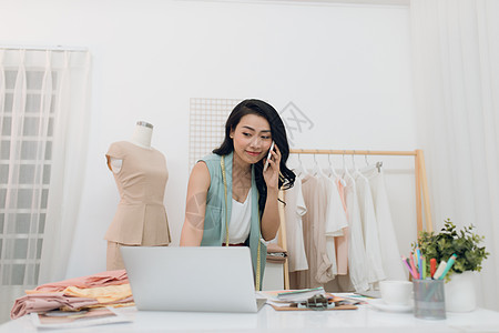 使用笔记本电脑和在工作室打电话的亚洲青年时装设计师商业女性互联网办公室企业家裁缝桌子生意模型纺织品衣服图片