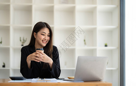 美丽的亚洲年轻女商务人士微笑着拿着咖啡杯和笔记本电脑在办公室工作 她笑着快乐女性商业商务成人享受热饮咖啡技术人士图片