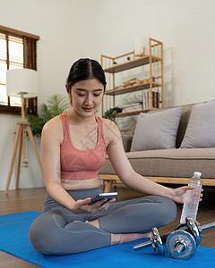 妇女坐在垫子上 拿着电话看网上健身训练的Tv课堂视频 在家使用健身应用程序女孩运动成人冥想瑜伽公寓女性运动员笔记本手机图片