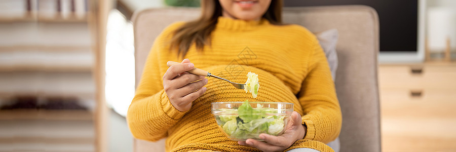 在家坐着吃沙拉的怀孕年轻孕妇快乐阅读母性饮食孩子们女性家庭早餐沙发微笑腹部图片