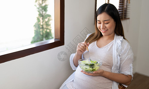 在家坐着吃沙拉的怀孕年轻孕妇快乐微笑腹部婴儿食物肚子水果饮食沙发女孩孩子们图片