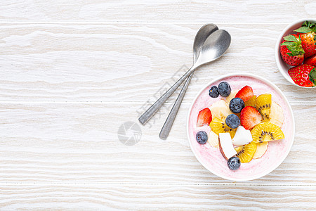 水果麦片健康早餐或甜点酸奶碗 配新鲜香蕉 草莓 蓝莓 可可 猕猴桃顶视图 带勺子的质朴木质白色背景 复制空间背景