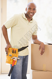 穿着工具带的男人在新家微笑时站在箱子旁站着房间房地产家具财产房子抵押阶梯男性相机购买者图片