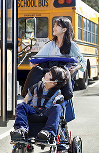 姐姐大姐在学校轮轮椅上推残疾兄弟孩子们友谊蓝色用具男生机动性学校公共汽车婴儿车防护图片