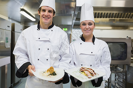 两个笑笑的厨师展示了盘子厨房烤箱烹饪男性食物柜台女士美食制服工作图片