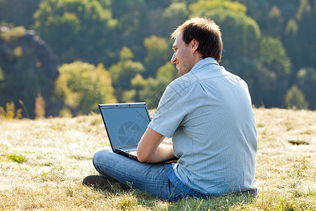 年轻人用笔记本电脑坐在山坡的草地上沉思自由太阳商业地平线城市风景全景技术微笑图片