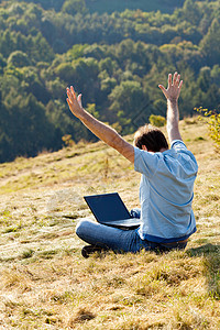 年轻人用笔记本电脑坐在山坡的草地上城市自由太阳技术商务沉思微笑场景天空风景图片